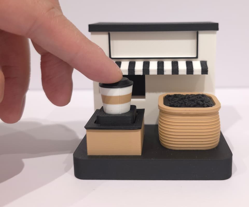 A finger pressing the takeaway cup on the printed coffee hatch, with its striped awning and sack of beans